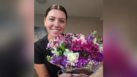 Natalie Dyson with brown hair in a pony tail and wearing gold earrings and black short sleeved top holding a bunch of white, purple and pink flowers hand tied towards the camera. She is standing in her kitchen. She is smiling.