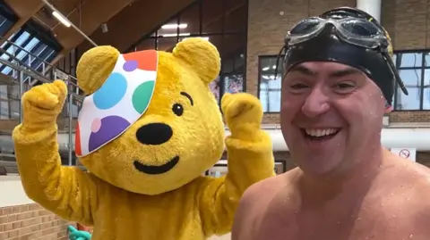 Pudsey, a human-sized yellow bear mascot wearing a patch over his eye, stands with his arms in the air. Next to him is Edward Sault, a topless man wearing goggles and a swimming cap.