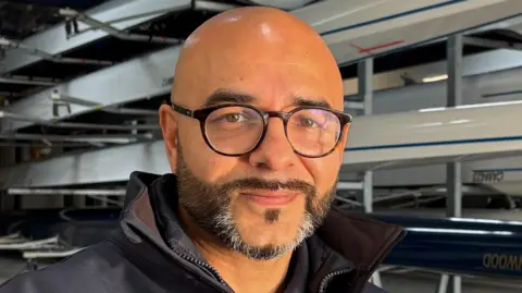 Omar Terywall, centre frame and looking directly out of the image. He is smiling slightly, has a dark, neatly trimmed beard, flecked with grey on his chin, and is wearing circular, black-framed glasses. He is bald. He is wearing a dark blue, collared top, but only the top of one shoulder on the left can be seen. Behind Omar are several sporting rowing boats mounted on racks inside a boathouse.