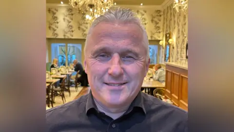 Roger Barrow Roger Barrow a man who is looking directly at the camera and smiling. He is standing up inside a tearoom and is wearing a black shirt.