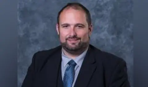 Worcestershire County Council A man with brown hair and a beard pictured in a black suit blazer, blue shirt and navy blue tie. He is slightly smiling and against a blue backdrop.