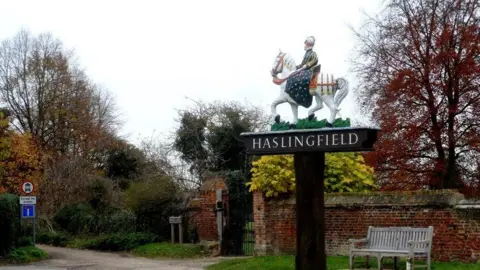 Bikeboy/Geograph Village sign for Haslingfield with a knight riding on a horse on top of the black-and-white sign.