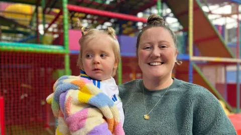 Lauren Byrne is smiling at the camera while holding her child Isla. They are in front of a colourful soft play area.