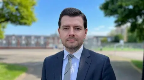Man in a blue, jacket, white shirt and grey tie looks at the camera. There is a school behind him.