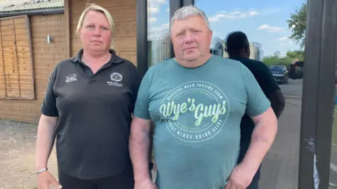 A woman with tied back blonde hair is wearing a black T-shirt and is stood in front of a wooden building. Next to her stands a man with grey hair wearing a green T-shirt with a large logo on.