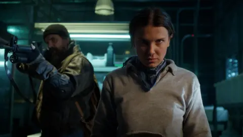 Netflix A still from a scene in season five of Stranger Things, with David Harbour poiting a rifle out of the frame, next to Millie Bobby Brown who stands with a focused expression and blood dribbling from one nostril