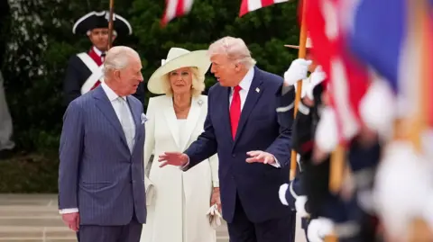 The King, Queen and Trump walk outside the White House with UK and US flags in the foreground.