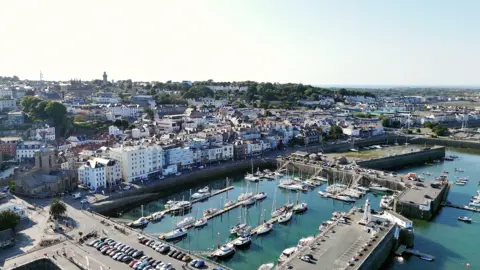 Aerial view of St Peter Port harbour in Guernsey, showing rows of white and pastel-coloured buildings along the waterfront, a marina filled with yachts and boats and green hills in the background under a clear blue sky. 