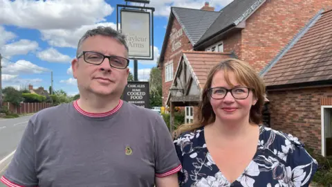 BBC The Hodsons stood side by side outside their pub. Mark is wearing a grey tshirt and Bonnie is wearing a dark blue dress with white flowers. The pub, behind them, is a large red brick building with 'The Tayleur Arms' painted on it in white writing. There is also a large freestanding pub sign with the pub's name on it. The sky is blue with white clouds. 