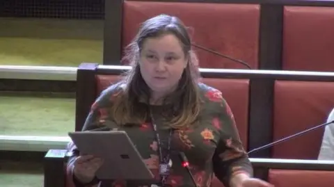 Warwickshire County Council Councillor Sarah Feeney, Labour opposition leader, stands during the 2025 Warwickshire County Council budget setting meeting. She is wearing a flowery top and holding a tablet computer. There are red leather and wooden benches behind her.