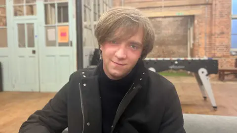 Josh McLaughlin/BBC Matthew Bothwell is sitting on a couch at the Spear Hastings youth hub. He is slightly tilting his head to his right while smiling for a photo. He is wearing a dark turtleneck and coat. A foosball table, wooden floors and brick walls form part of a blurred background.