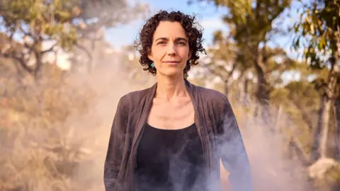 Nic Vevers A woman, with short curly brown hair and wearing a dark top, stands in an open forest area with wisps of smoke surrounding her