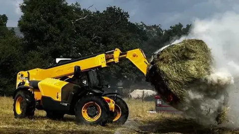 DWFRS A yellow telehandler holds a smouldering hay bale aloft in a field with smoke billowing.