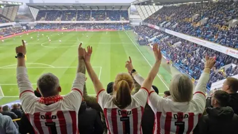 Kirsty Cummings Three people are at a game wearing Sunderland tops with their arms in the air. They all have the word Dad on the back of their tops. 