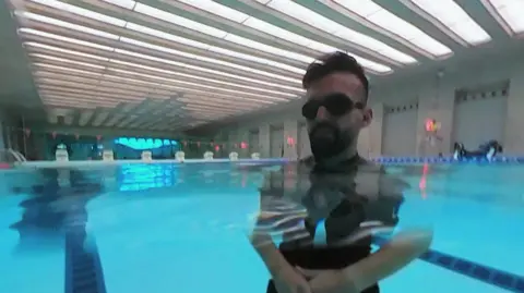A man is standing in a swimming pool. He is wearing googles.