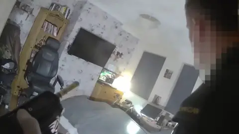 PA Media A paramedic in green uniform is seen to the right, as a camera behind captures him entering a room with white walls, bed and TV mounted on the wall. Computer monitors can be seen on drawers and a desk and an office chair against the wall. A grey duvet covers the bed.