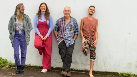 Heidi Burton Andrew stands against a white wall outside, with three members of his band The Promise. They are all wearing colourful clothes and smiling at the camera.