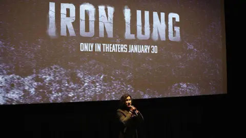 Getty Mark aparece con un traje negro, guantes marrones y un micrófono frente a una pantalla de cine.