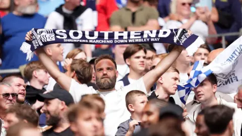 A close up shot of part of the mostly male crowd at a football match. A man holds aloft a navy and white scarf with the words "SOUTHEND. SHRIMPERS MARCHING DOWN WEMBLEY WAY"