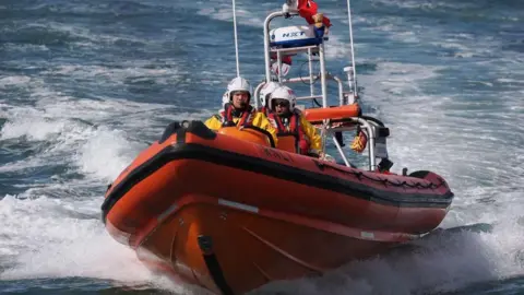 Close up shot of a lifeboat and its crew. The boat is bright orange and there are four crew members on board, wearing yellow overalls, lifejackets and white helmets.