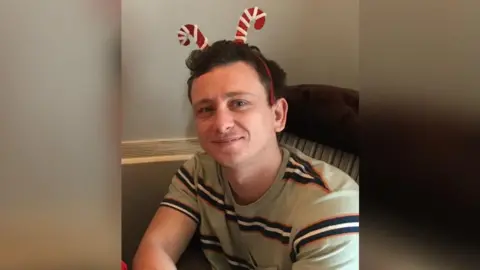 Sara Foster A man with short brown hair is smiling and wearing a candy cane headband and a beige top with black and white stripes. He is sitting indoors on a chair.