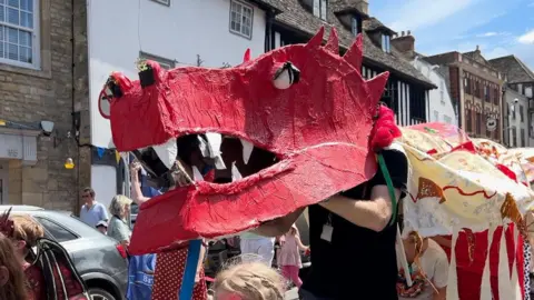 A large red dragon's head, carried by adults and children, is seen being carried down a picturesque high street. The cloth 'body' of the dragon can be seen behind carried by other children.