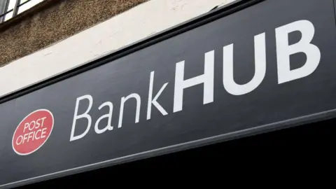 Getty Images A close-up of a sign for a Post Office-run banking hub. The black sign sits above the entrance and says "Bank Hub" in white writing. A red oval Post Office logo sits to the left of the writing