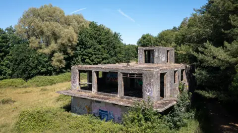 A derelict concrete building of two storeys plus a small third storey room sits on the edge of woodland.
