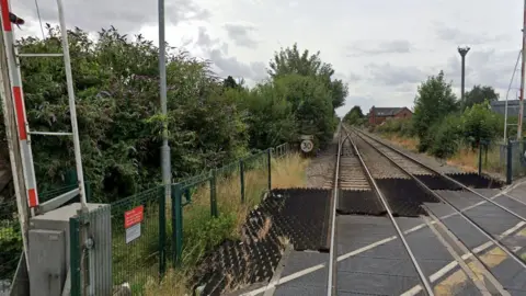 Two sets of train tracks on a level crossing in Winsover Road, which is flanked by large bushes and trees. There is a red and white barrier on the left in the upright position.