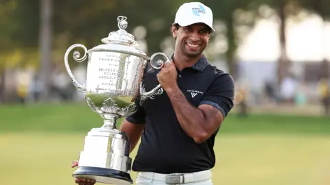 Reuters Aaron Rai holding the The Wanamaker Trophy and smiling. he is wearing a black t-shirt and a white cap