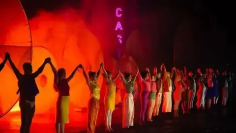 Cast A group of theatre performers stand in a line and hold hands aloft on a stage with misty orange lighting