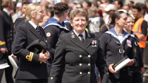 An image of a group of Royal Navy sailors marching at London's pride parade in 2024. In the foreground is an older female, in uniform and wearing medals, while two younger sailors behind her look to the people lining the street.