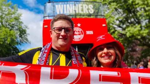 A man and woman in aberdeen hats, holding a scarf out in front of a bus.
