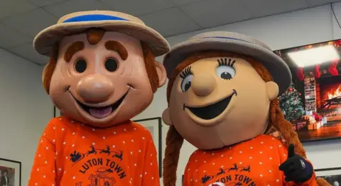Roger Wash Luton Town mascots Happy Harry and Happy Hatty wearing orange jumpers and straw hats. They have large, cartoon-like faces.