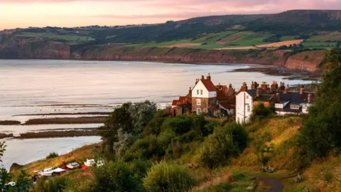 Getty Images A general view of Robin Hood's Bay in a soft pink light either at dawn or dusk. The bay curves round, with some small finishing boats and houses in the foreground, to a more rugged part further along, The tide is almost completely in, leaving only small patched of beach exposed.