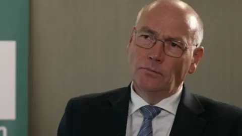Adrian Usher is a middle-aged man, largely bald but with some short white hair at the back of his head and around his temples. He has brown eyes and is wearing glasses, a blue tie, white shirt and black suit jacket. He's in what looks like a basic office, with a beige wall behind him, and has a serious expression.