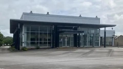 Former Mercedes car dealership building with glass doors and a concrete yard outside.
