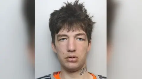 Robert Leverton pictured staring straight ahead. He has dark brown hair and is wearing an orange t-shirt underneath a grey t-shirt. He has spots on his face and lips and a tattoo on his neck.