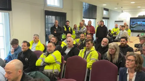 Rows of people - largely men in high vis coats, sitting in a council meeting room with others standing behind them and looking towards the front