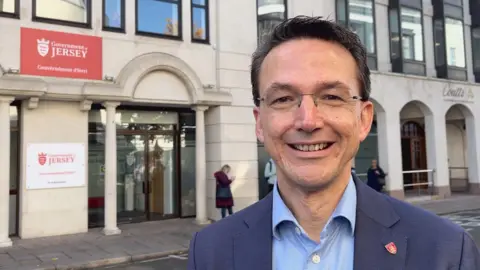 A man in a suit smiling at the camera in front of government offices.