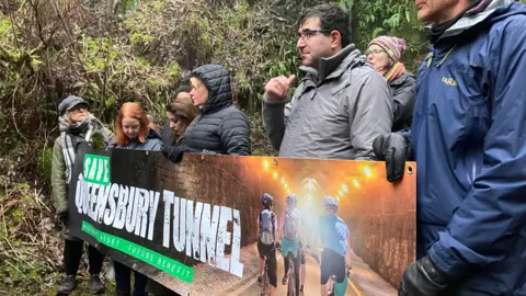 Olivia Richwald/BBC A photo taken from the side of ten or so people standing behind a banner reading "save Queensbury tunnel".