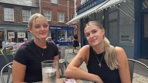 Maisy Collins, with tied back blond hair and a black t shirt, sits at an outside table next to Abbie Davis, with long blond hair and a black vest top. Miss Davis also has two tattoos, one on each arm. The cafe they work at is visible in the background and a glass of water is on the table in front of them.