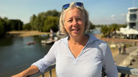 A woman with short blonde hair stands in front of a harbour smiling into the camera. It is a sunny day.