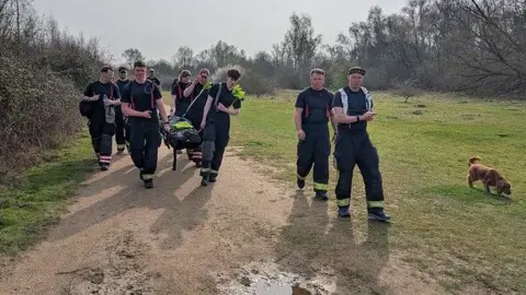 A group of nine young people, wearing dark coloured t-shirts and trousers with fluorescent stripes towards the bottom walking along a path towards the camera. Some of them are holding up a stretcher with a dummy lying on it. Around them is grass and trees. There is also a small dog on the grass. 