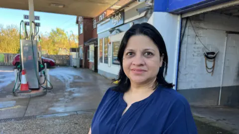 A head and shoulders shot of a woman with black hair, wearing a dark blue blouse. She stands on the forecourt of a petrol station, with pumps, a shop and a garage area in the background.