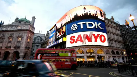 Digital billboards lit up on a building in Piccadilly Circus in London. There is a red double decker bus passing beneath the billboard and a black taxi. A crowd is on the pavement beneath the billboard. 