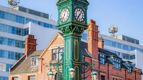 Birmingham Heritage Week A green clock tower in front of a red brick building with two grey office blocks directly behind