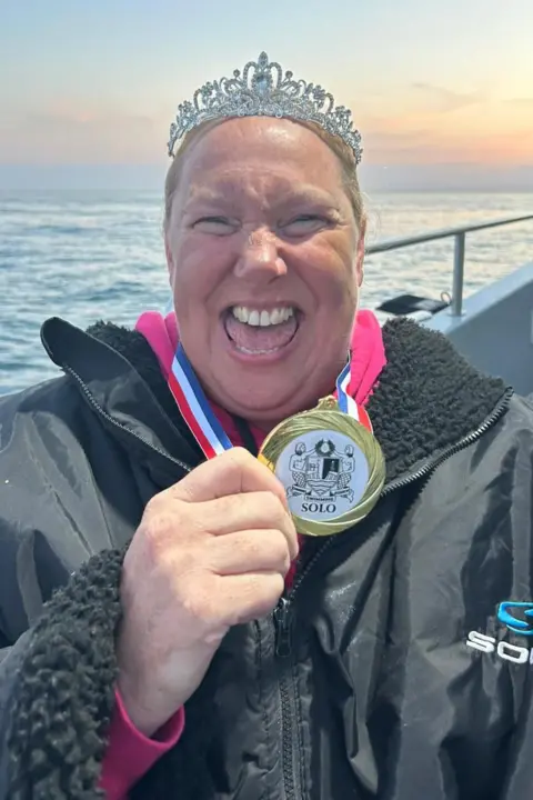 Lindsey Walke Lindsey Walke, wearing a swim robe and a tiara, she is holding her medal. She is facing the camera and smiling with her eyes squinted. Behind her is the side of the boat. The sea behind her is a light blue and the sky is a light blue to orange ombre.
