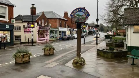 Amy Holmes/BBC A quiet small‑town high street on a grey day, with independent shops lining the road and a decorative “Woburn Sands” sign standing in the foreground beside planters and paved walkways.