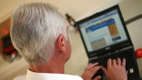 PA Media A grey-haired man is sat at a computer. The back of his head can be seen and his hands on the laptop keyboard. The laptop is in the background and the screen cannot be seen clearly but shows what appears to be the layout of a webpage.